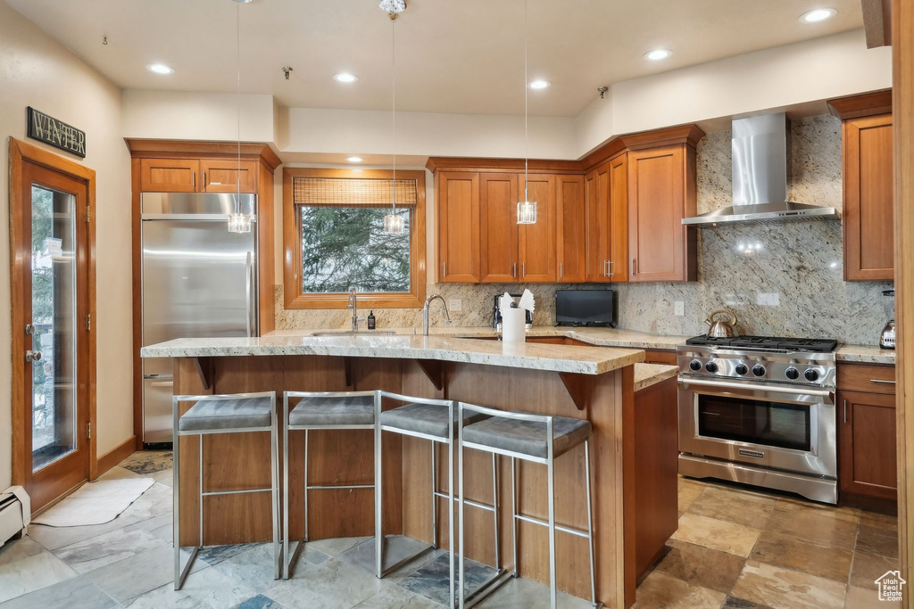 Kitchen featuring wall chimney exhaust hood, light stone countertops, high quality appliances, a sink, and brown cabinets