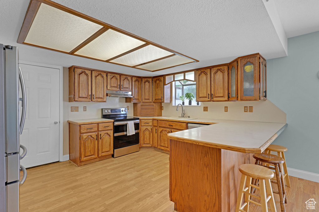 Kitchen featuring brown cabinetry, a peninsula, light wood-style floors, under cabinet range hood, and stainless steel appliances