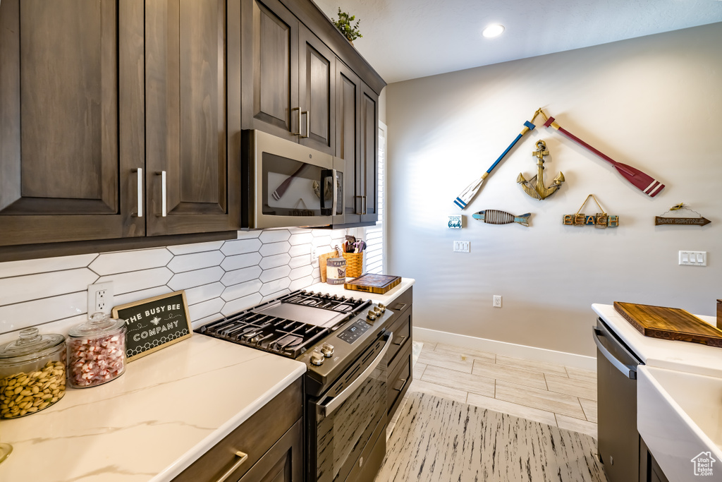 Kitchen featuring decorative backsplash, dark brown cabinets, light stone countertops, baseboards, and appliances with stainless steel finishes