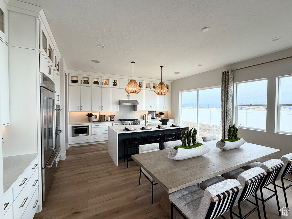 Kitchen featuring decorative backsplash, a center island with sink, light countertops, under cabinet range hood, and dark wood finished floors