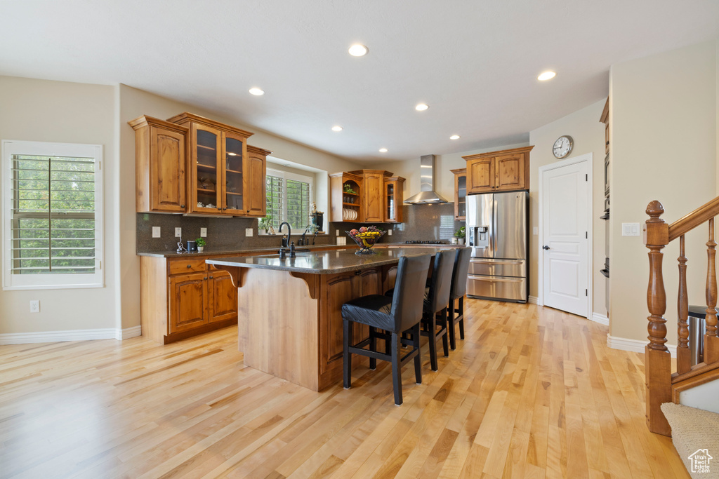 Kitchen featuring tasteful backsplash, a breakfast bar, a kitchen island with sink, stainless steel refrigerator with ice dispenser, and wall chimney exhaust hood