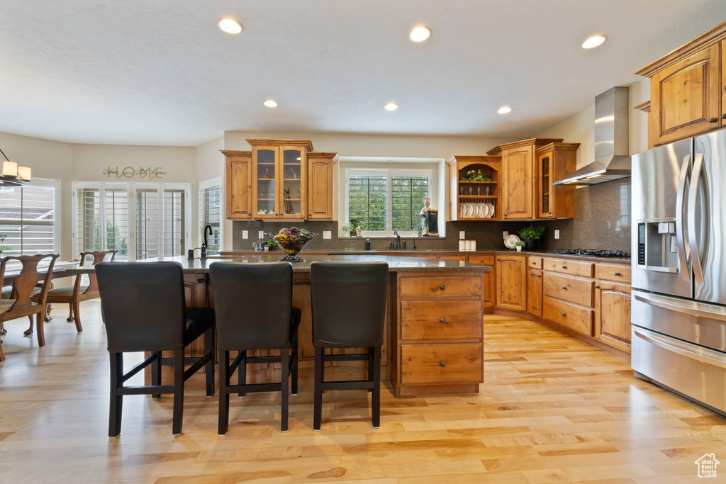 Kitchen featuring a breakfast bar, wall chimney exhaust hood, brown cabinetry, stainless steel fridge with ice dispenser, and a kitchen island