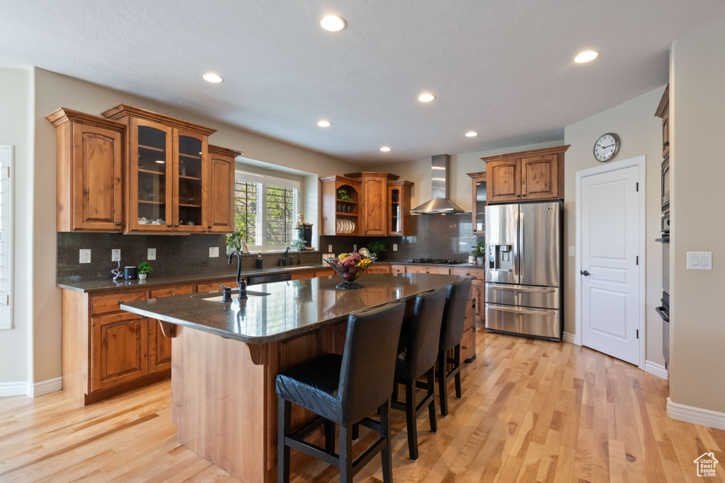 Kitchen with brown cabinets, appliances with stainless steel finishes, a sink, a kitchen island with sink, and wall chimney exhaust hood