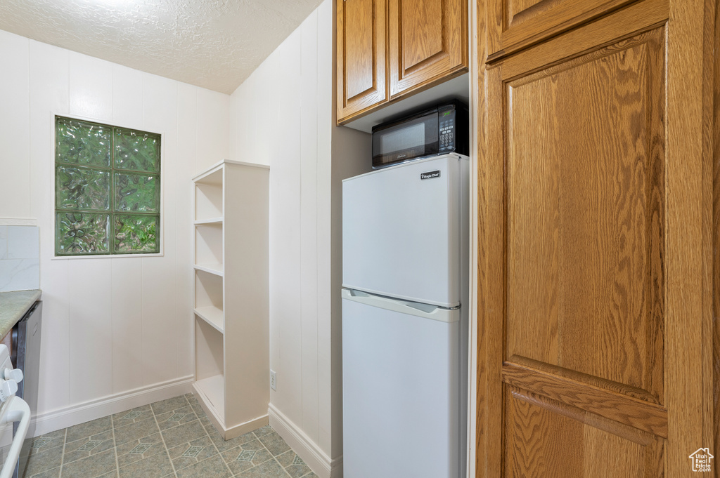 Kitchen featuring brown cabinetry, freestanding refrigerator, black microwave, baseboards, and a textured ceiling