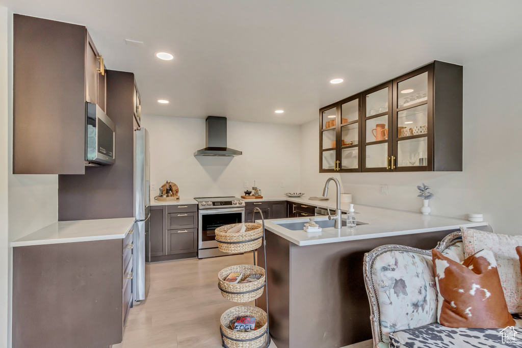 Kitchen with appliances with stainless steel finishes, a sink, a peninsula, glass insert cabinets, and wall chimney exhaust hood