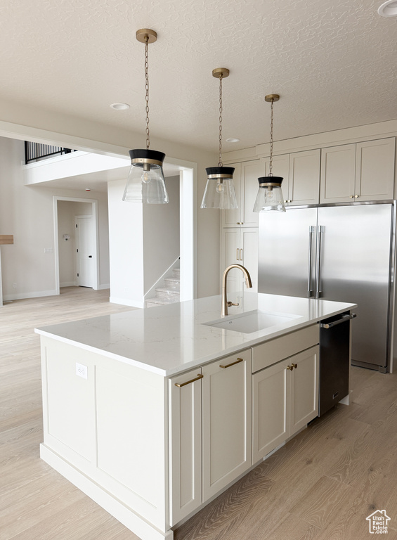 Kitchen featuring high end refrigerator, a sink, an island with sink, light wood-type flooring, and light stone countertops
