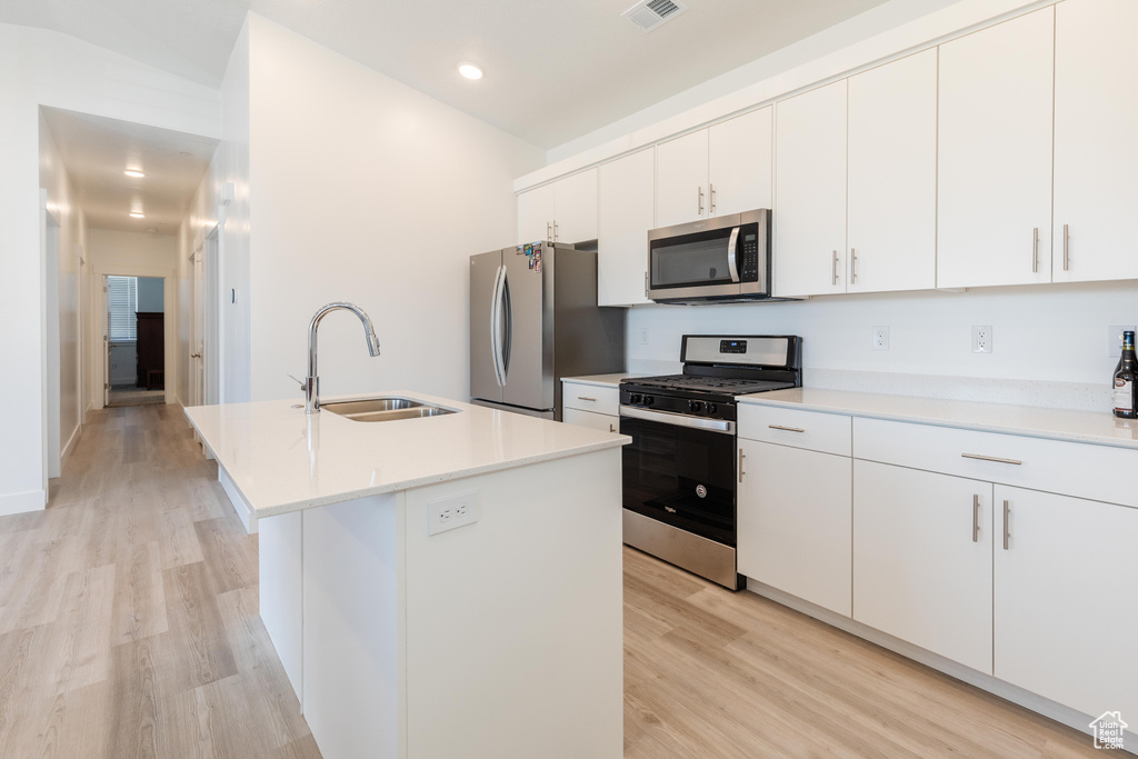 Kitchen featuring appliances with stainless steel finishes, a sink, light wood finished floors, light countertops, and a center island with sink