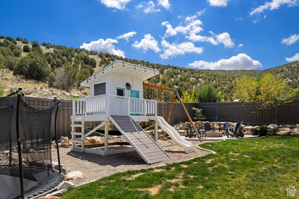 View of playground with a fenced backyard, a lawn, a trampoline, an outdoor fire pit, and a forest view