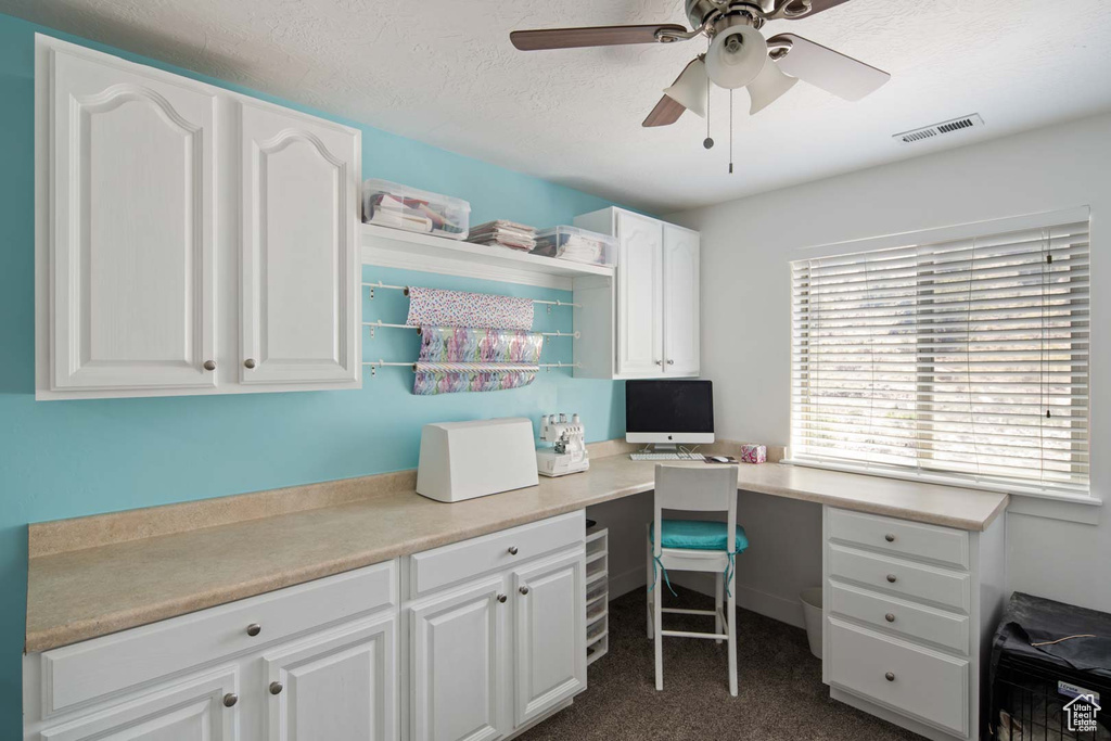 Office space featuring ceiling fan, a textured ceiling, visible vents, built in desk, and dark carpet