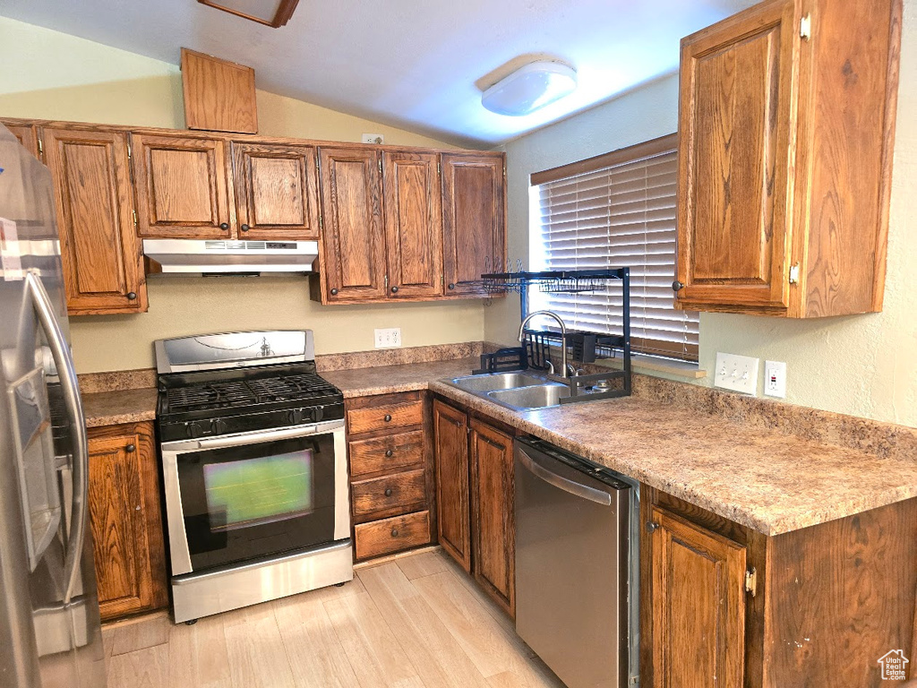 Kitchen with appliances with stainless steel finishes, brown cabinetry, light wood-type flooring, under cabinet range hood, and vaulted ceiling