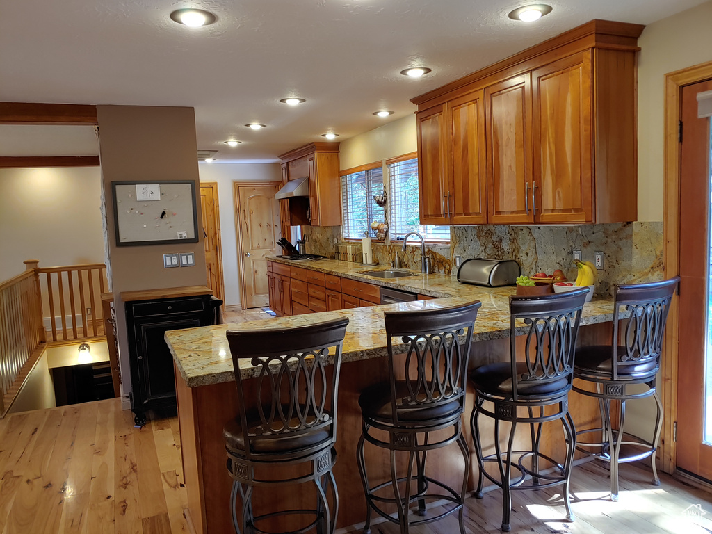 Kitchen featuring brown cabinetry, backsplash, a sink, light stone counters, and ventilation hood
