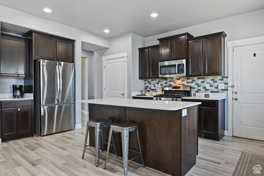 Kitchen with dark brown cabinets, light wood-type flooring, light countertops, backsplash, and stainless steel appliances