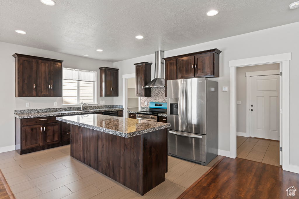 Kitchen with a kitchen island, appliances with stainless steel finishes, dark brown cabinets, light wood finished floors, and wall chimney exhaust hood