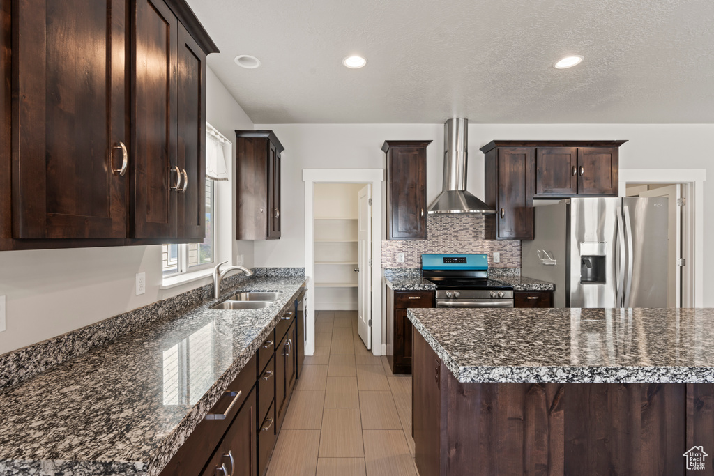 Kitchen with wall chimney range hood, dark brown cabinets, a sink, stainless steel appliances, and decorative backsplash