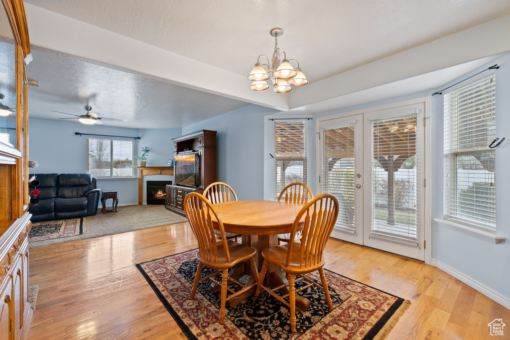 Dining space with light wood-style floors, baseboards, a lit fireplace, french doors, and ceiling fan with notable chandelier