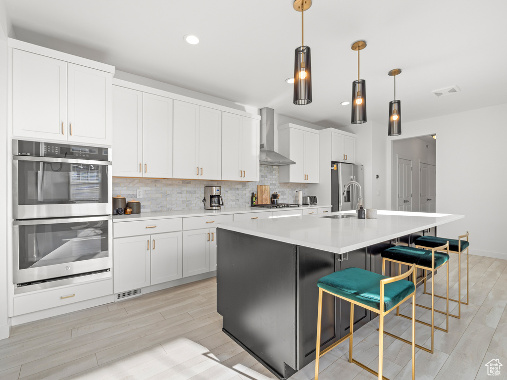 Kitchen featuring light wood-type flooring, wall chimney range hood, stainless steel appliances, a center island with sink, and backsplash