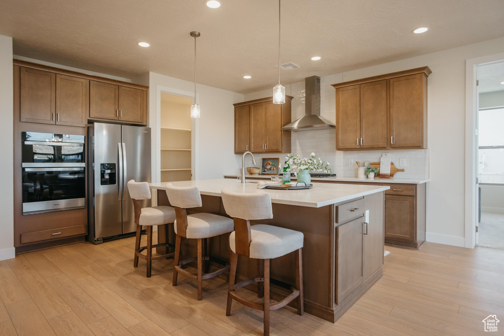 Kitchen with light wood-style flooring, wall chimney exhaust hood, a center island with sink, and stainless steel appliances
