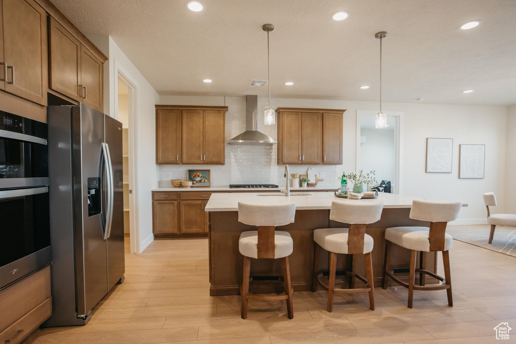 Kitchen featuring brown cabinets, light wood finished floors, wall chimney exhaust hood, recessed lighting, and stainless steel refrigerator with ice dispenser