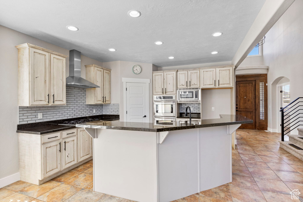 Kitchen with wall chimney range hood, appliances with stainless steel finishes, a kitchen island with sink, baseboards, and tasteful backsplash