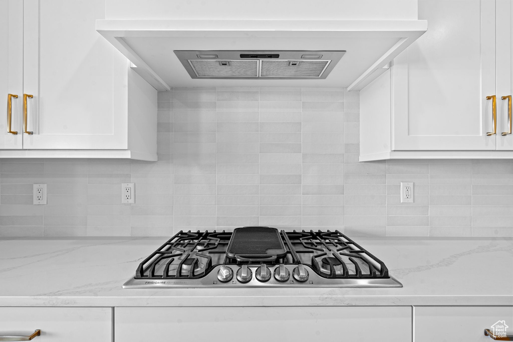 Kitchen with light stone counters, ventilation hood, stainless steel gas cooktop, white cabinetry, and backsplash