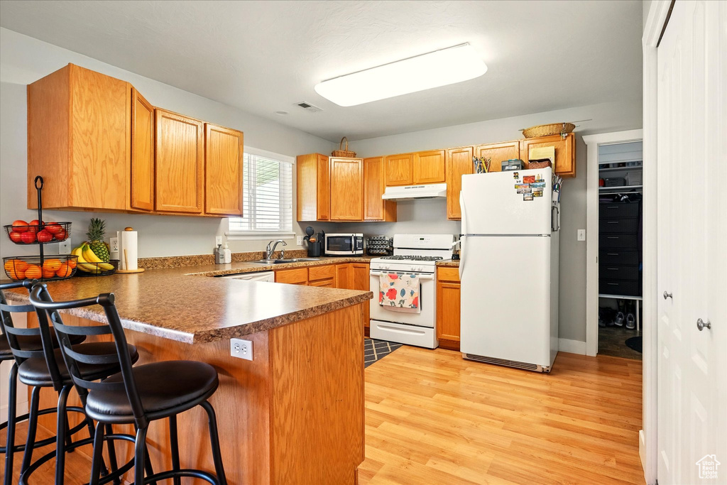 Kitchen with a sink, a peninsula, under cabinet range hood, white appliances, and light wood-style flooring