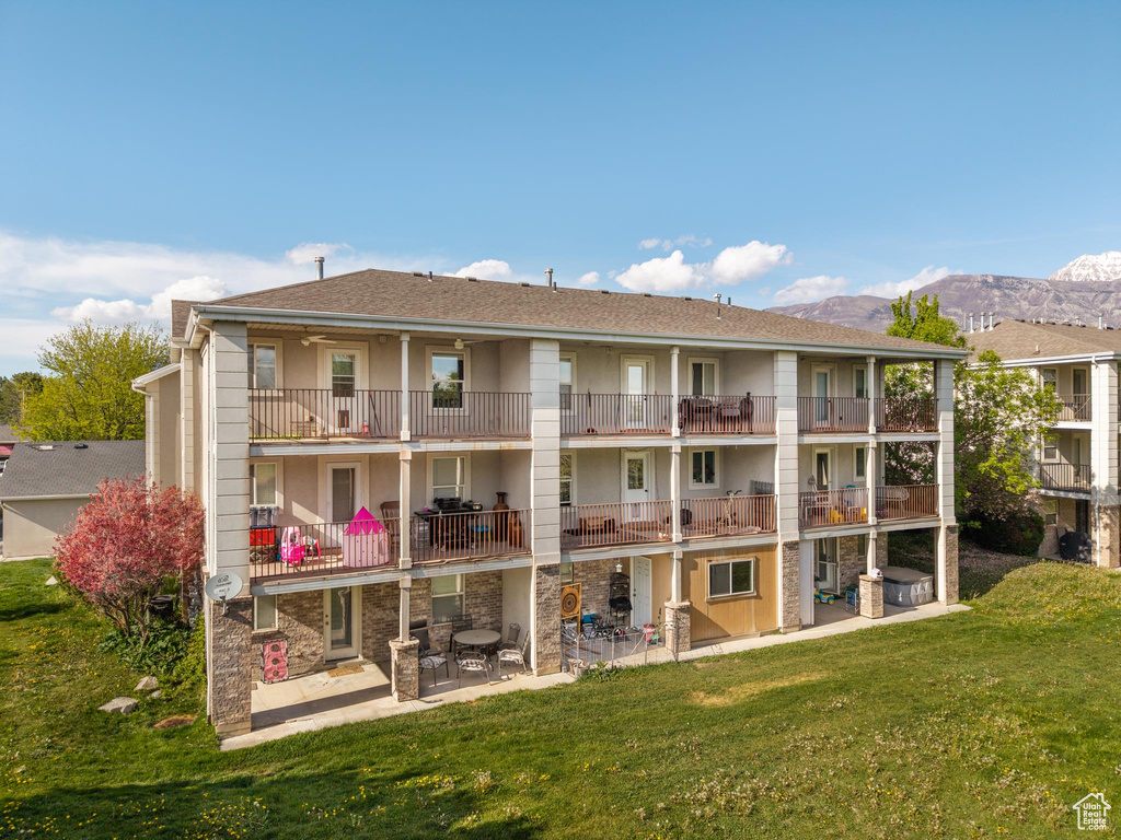 Back of house featuring a shingled roof, stucco siding, a mountain view, a patio area, and a lawn