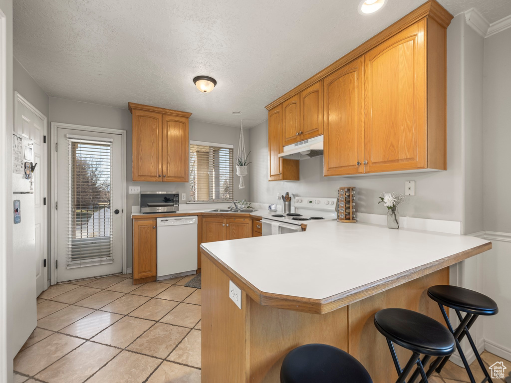 Kitchen featuring a peninsula, white appliances, a kitchen bar, under cabinet range hood, and a sink