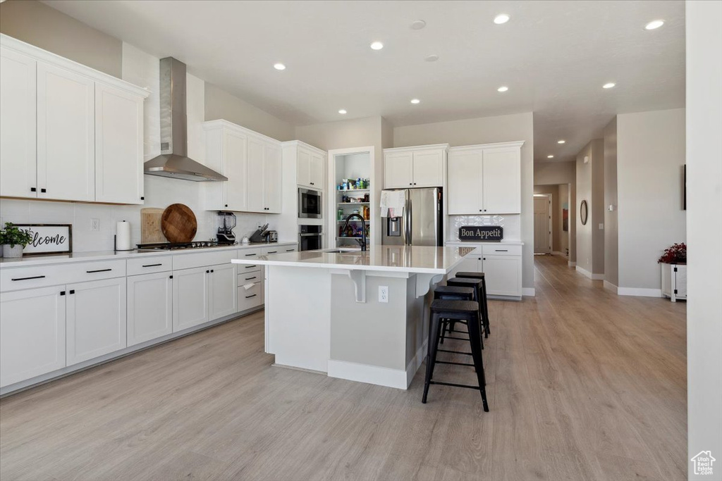 Kitchen featuring light wood-type flooring, wall chimney exhaust hood, a sink, appliances with stainless steel finishes, and backsplash