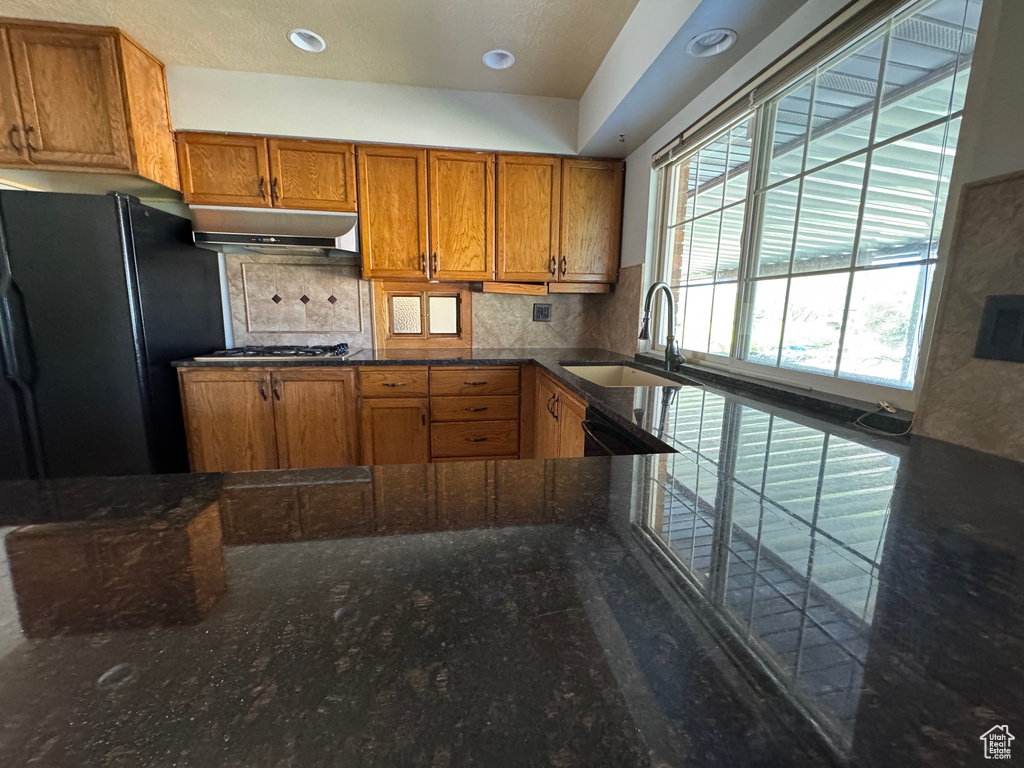 Kitchen featuring freestanding refrigerator, under cabinet range hood, a sink, backsplash, and brown cabinets