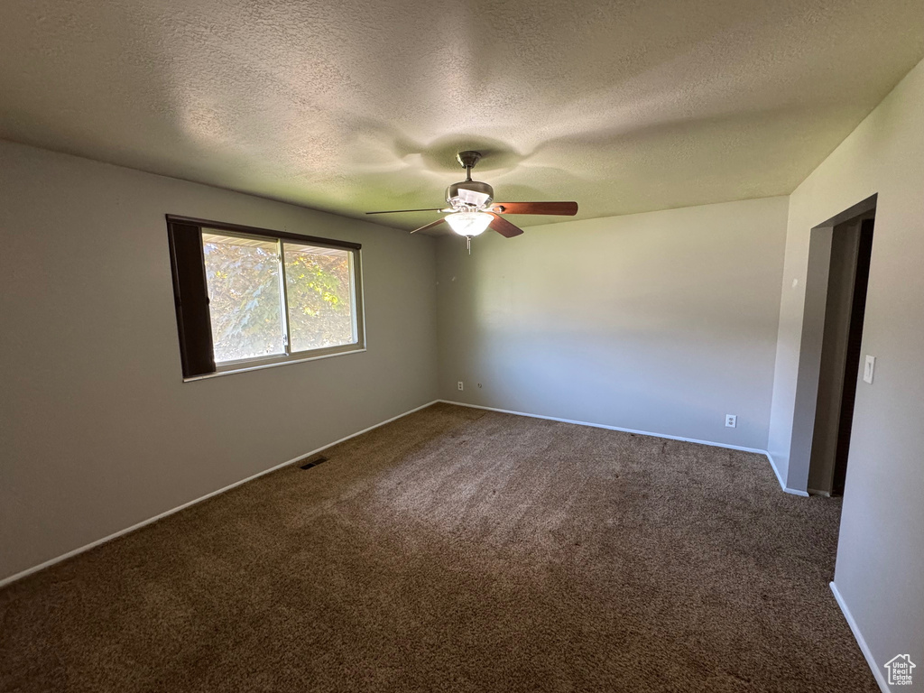 Empty room with a textured ceiling, dark colored carpet, a ceiling fan, and visible vents