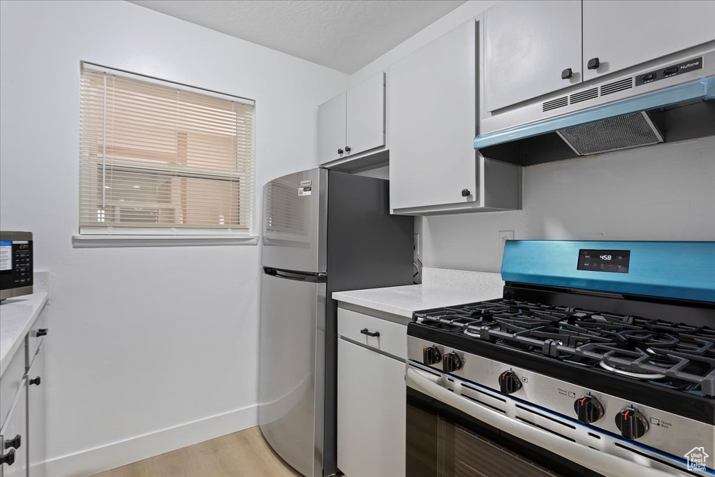 Kitchen with stainless steel appliances, under cabinet range hood, light countertops, white cabinetry, and light wood finished floors
