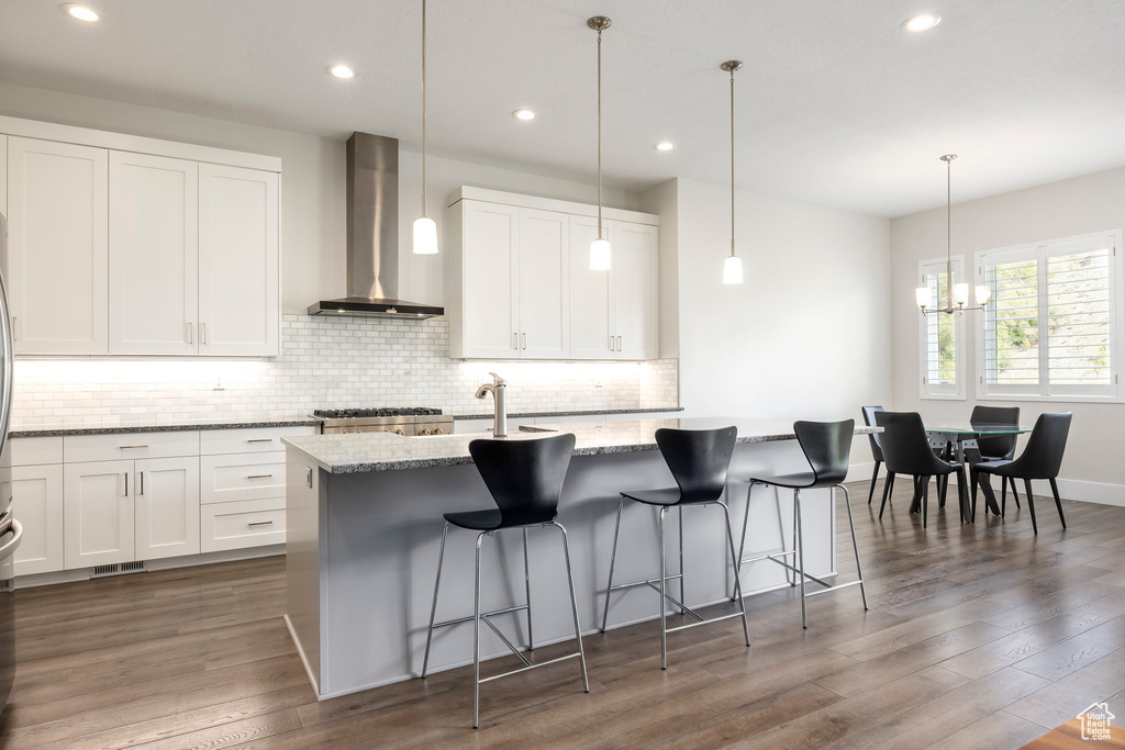 Kitchen featuring light stone counters, wall chimney exhaust hood, dark wood-type flooring, backsplash, and a kitchen island with sink