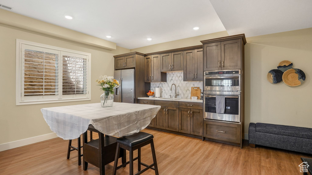 Kitchen featuring dark brown cabinetry, appliances with stainless steel finishes, decorative backsplash, and light wood-style flooring