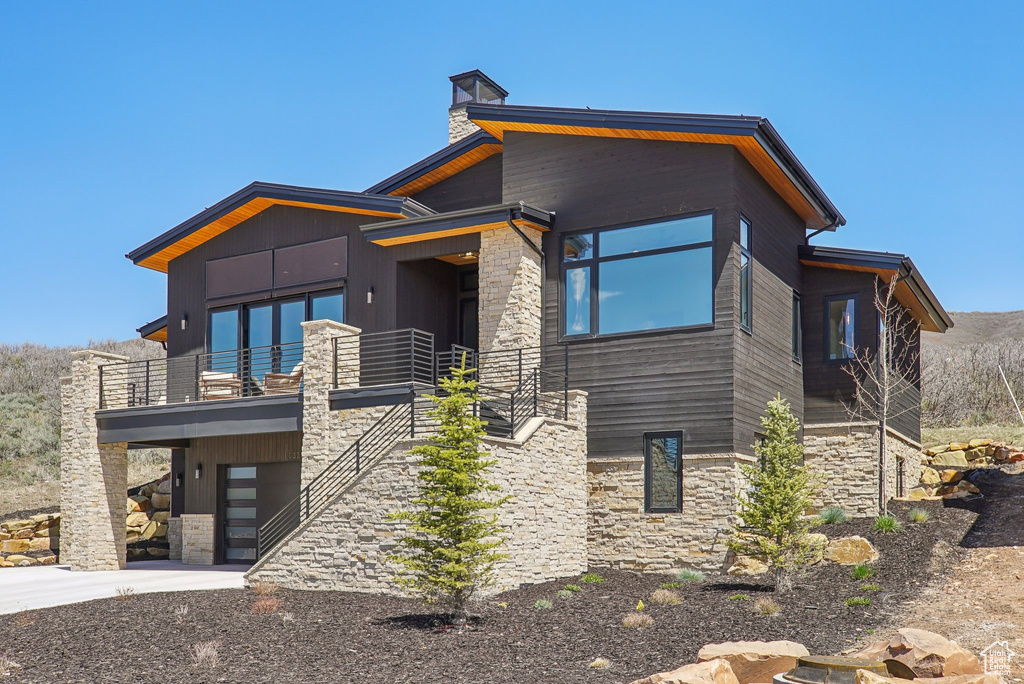 Contemporary home with an attached garage, stone siding, driveway, a balcony, and a chimney