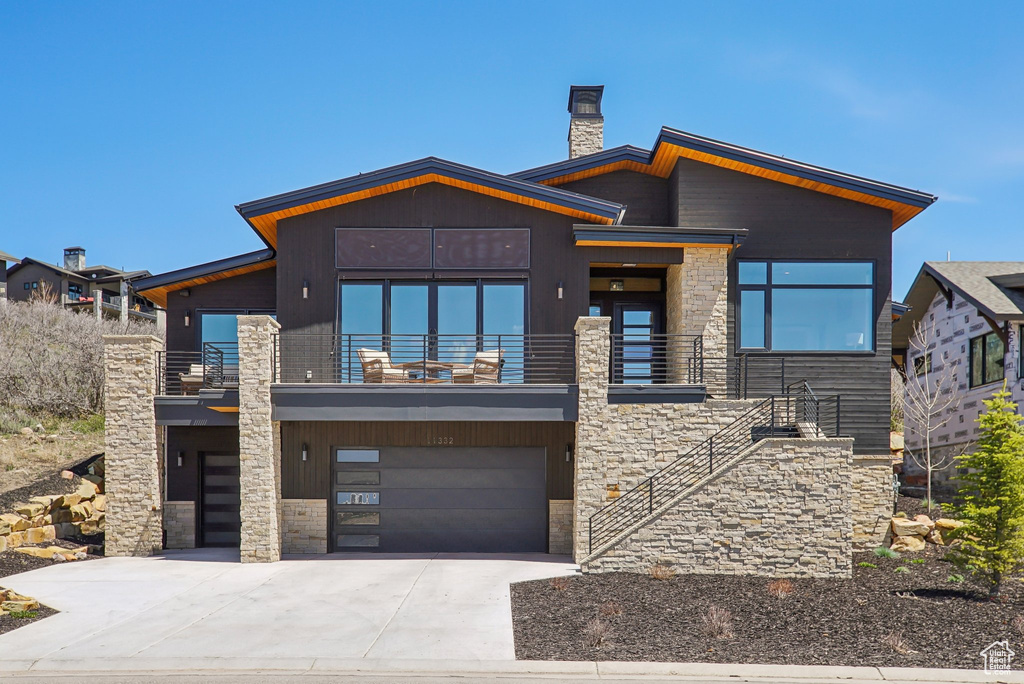Modern home featuring a balcony, a chimney, driveway, an attached garage, and stone siding