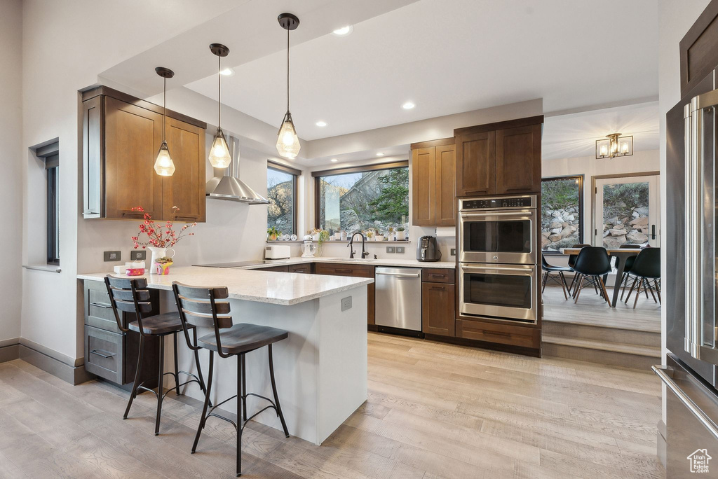 Kitchen featuring a sink, a peninsula, wall chimney exhaust hood, stainless steel appliances, and light wood-type flooring