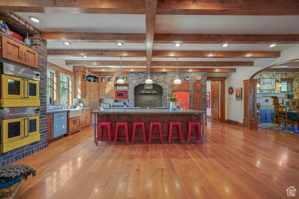Kitchen featuring stainless steel double oven, light wood-type flooring, a kitchen breakfast bar, wall chimney exhaust hood, and beam ceiling