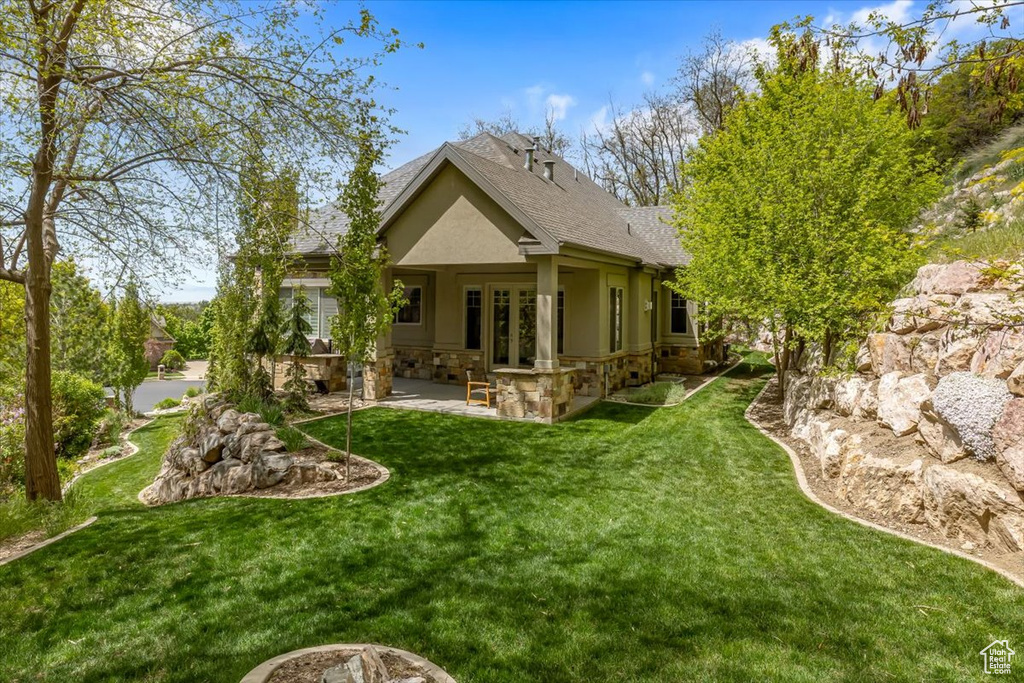 Back of house with stone siding, a patio, stucco siding, a yard, and roof with shingles