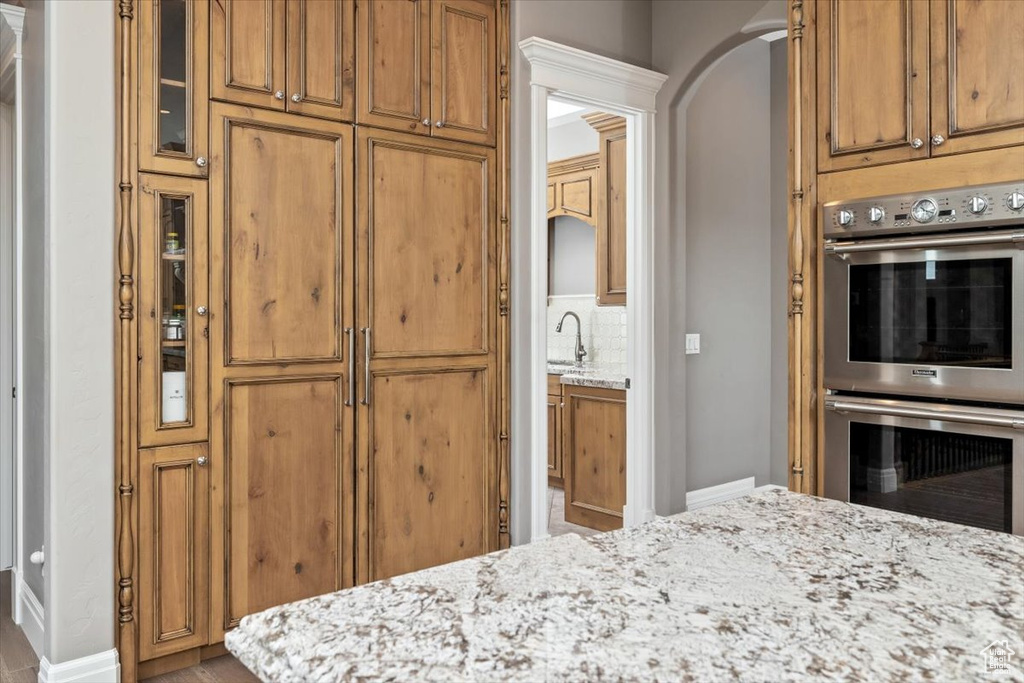 Kitchen with brown cabinetry, stainless steel double oven, baseboards, and light stone counters
