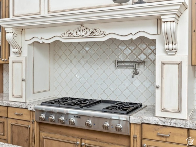 Kitchen with brown cabinets, stainless steel gas stovetop, light wood-type flooring, backsplash, and light stone countertops