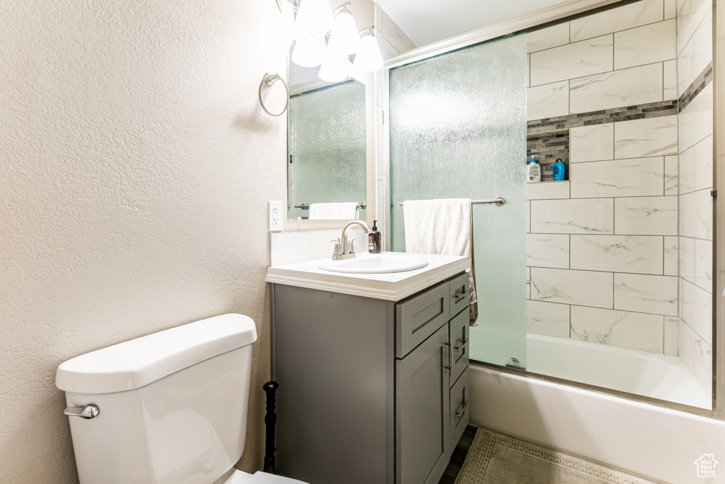 Bathroom with toilet, vanity, combined bath / shower with glass door, and a textured wall