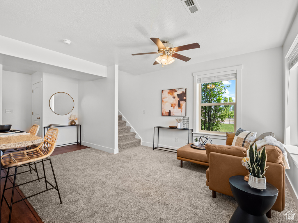 Carpeted living room with ceiling fan, visible vents, baseboards, and stairway