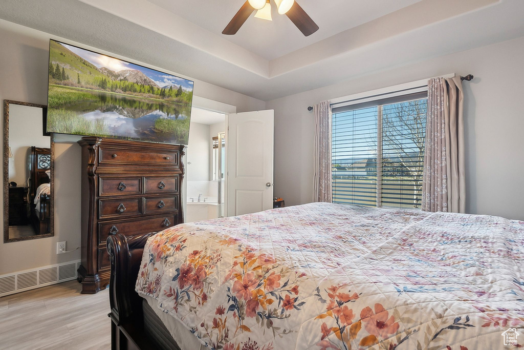 Bedroom featuring light wood finished floors, a ceiling fan, a tray ceiling, visible vents, and ensuite bath