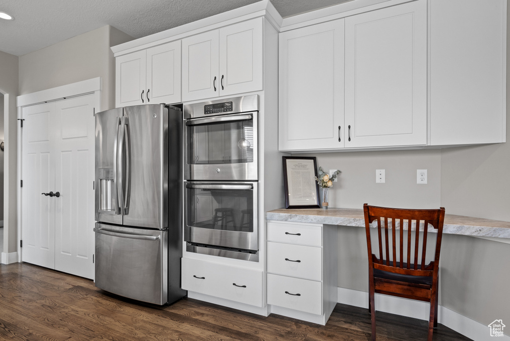 Kitchen featuring built in desk, stainless steel appliances, white cabinetry, dark wood-type flooring, and baseboards