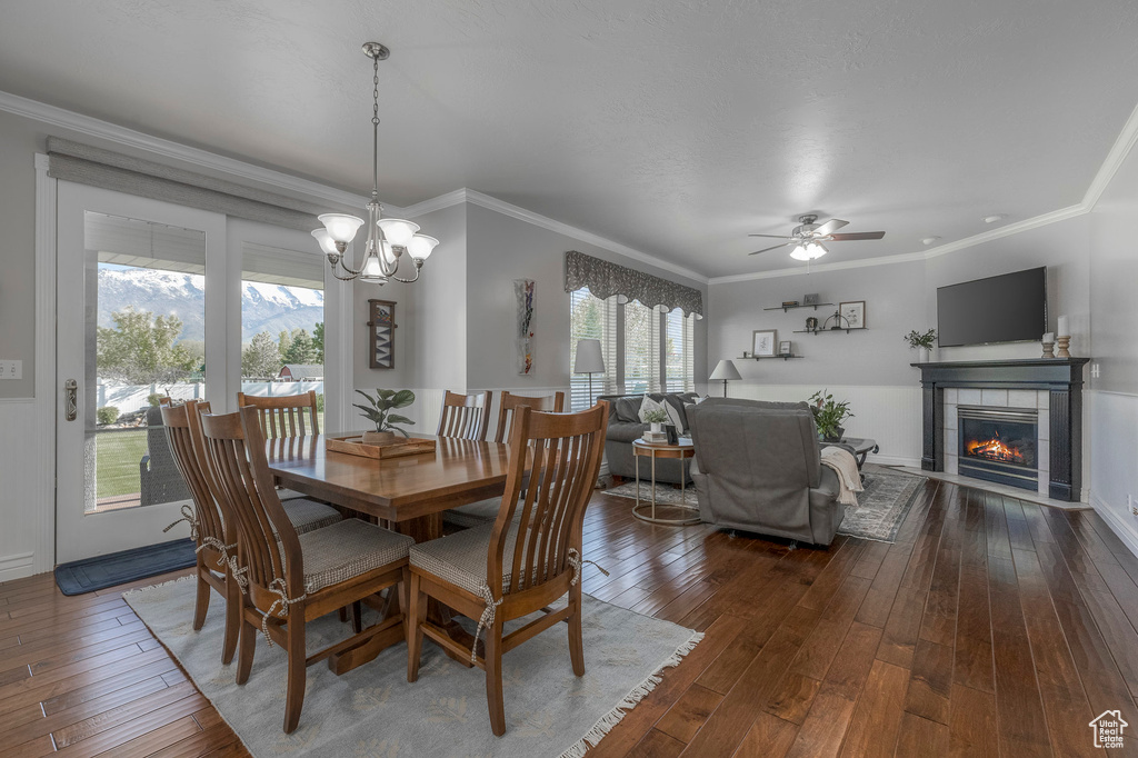 Dining room featuring crown molding, ceiling fan with notable chandelier, dark wood-style floors, and a tiled fireplace