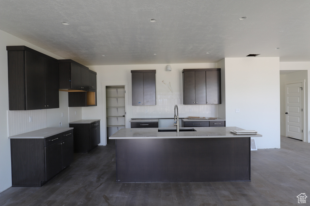 Kitchen featuring a sink, a center island with sink, light countertops, tasteful backsplash, and dark wood-style floors