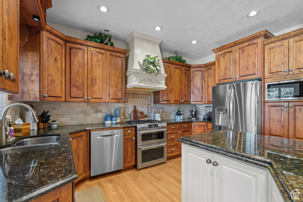 Kitchen featuring dark stone counters, custom range hood, a sink, light wood-style flooring, and stainless steel appliances