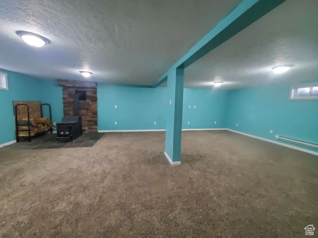 Basement with carpet, a textured ceiling, a baseboard heating unit, baseboards, and a wood stove