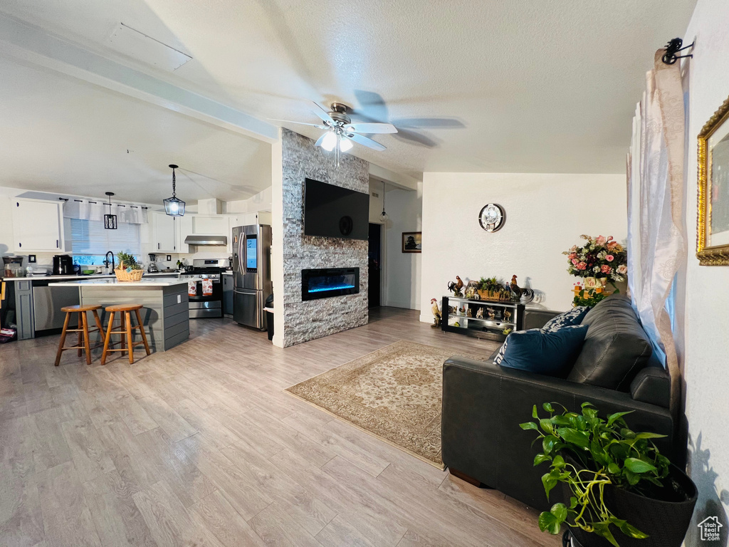 Living area with a stone fireplace, light wood-type flooring, lofted ceiling with beams, and ceiling fan