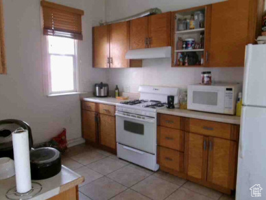 Kitchen featuring brown cabinets, light countertops, light tile patterned floors, under cabinet range hood, and white appliances